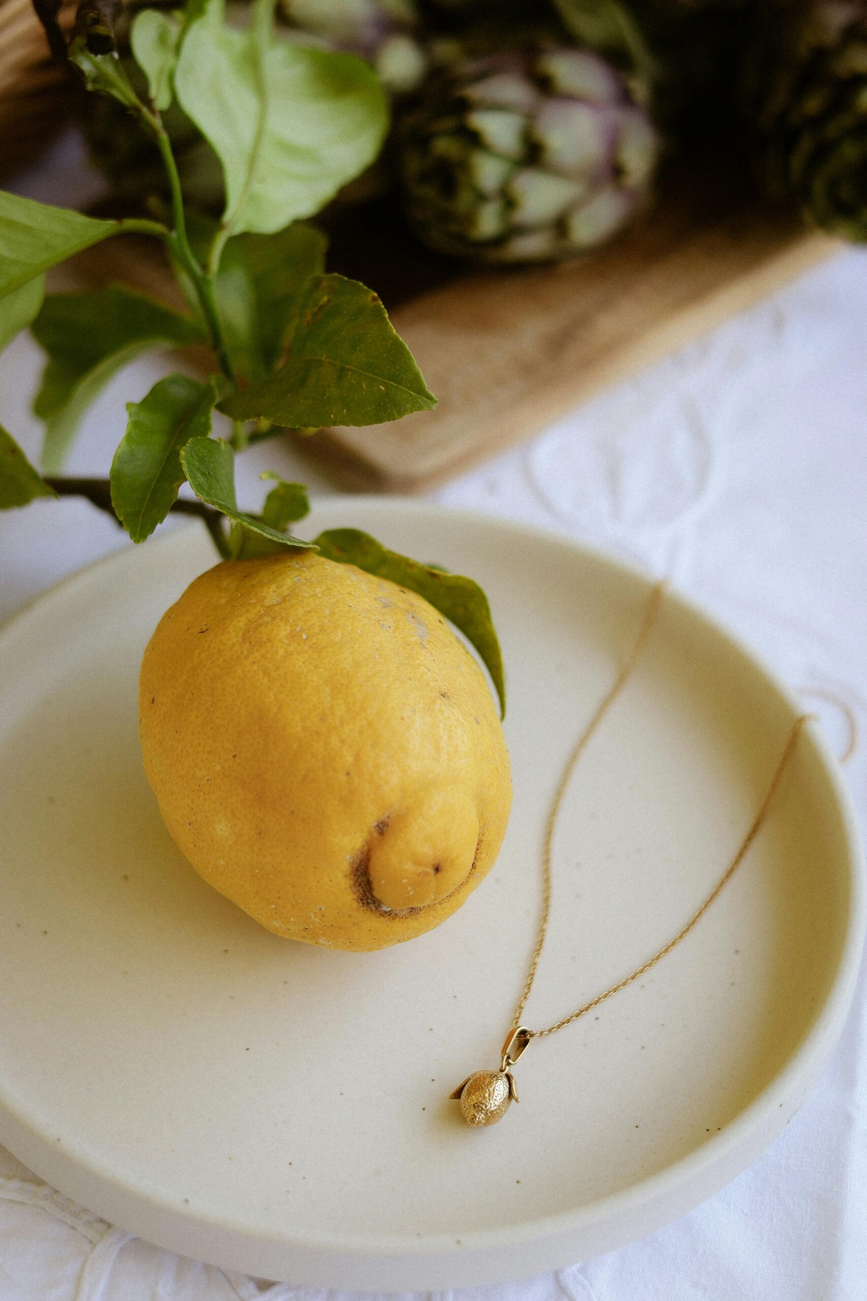 Lemon with gold necklace on plate, artichokes in soft focus. Elegant still life composition.